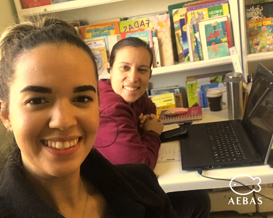 duas mulheres sorrindo. Ao fundo estante de livros infantis.